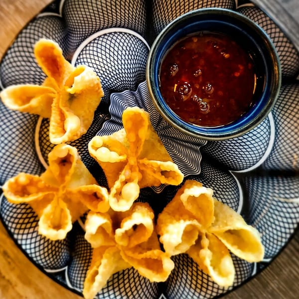 Golden-fried crab rangoons arranged on a patterned plate with a side of sweet chili dipping sauce.