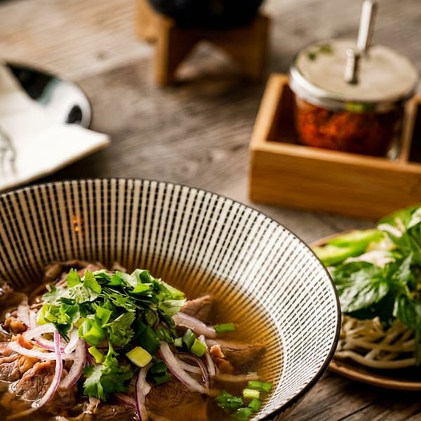 A bowl of pho with beef, onions, and cilantro, with a side of fresh herbs.