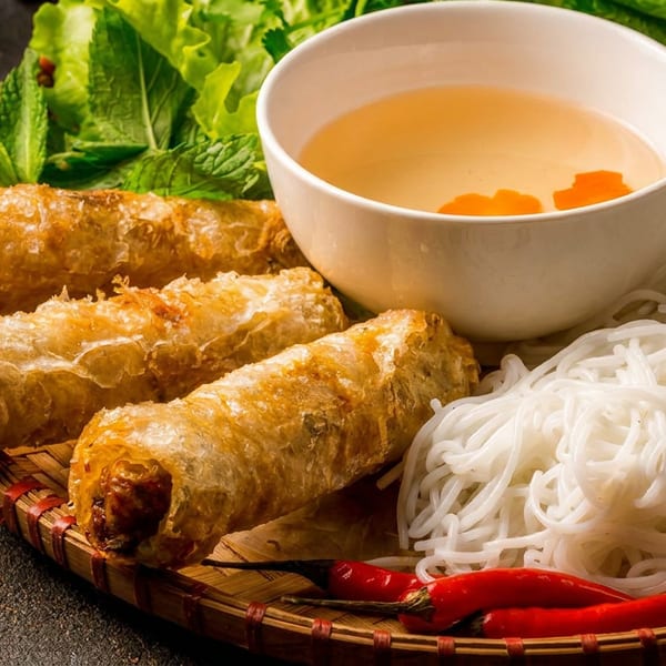 Crispy fried spring rolls, rice vermicelli, fresh herbs, cucumber, and dipping sauce on a woven tray.